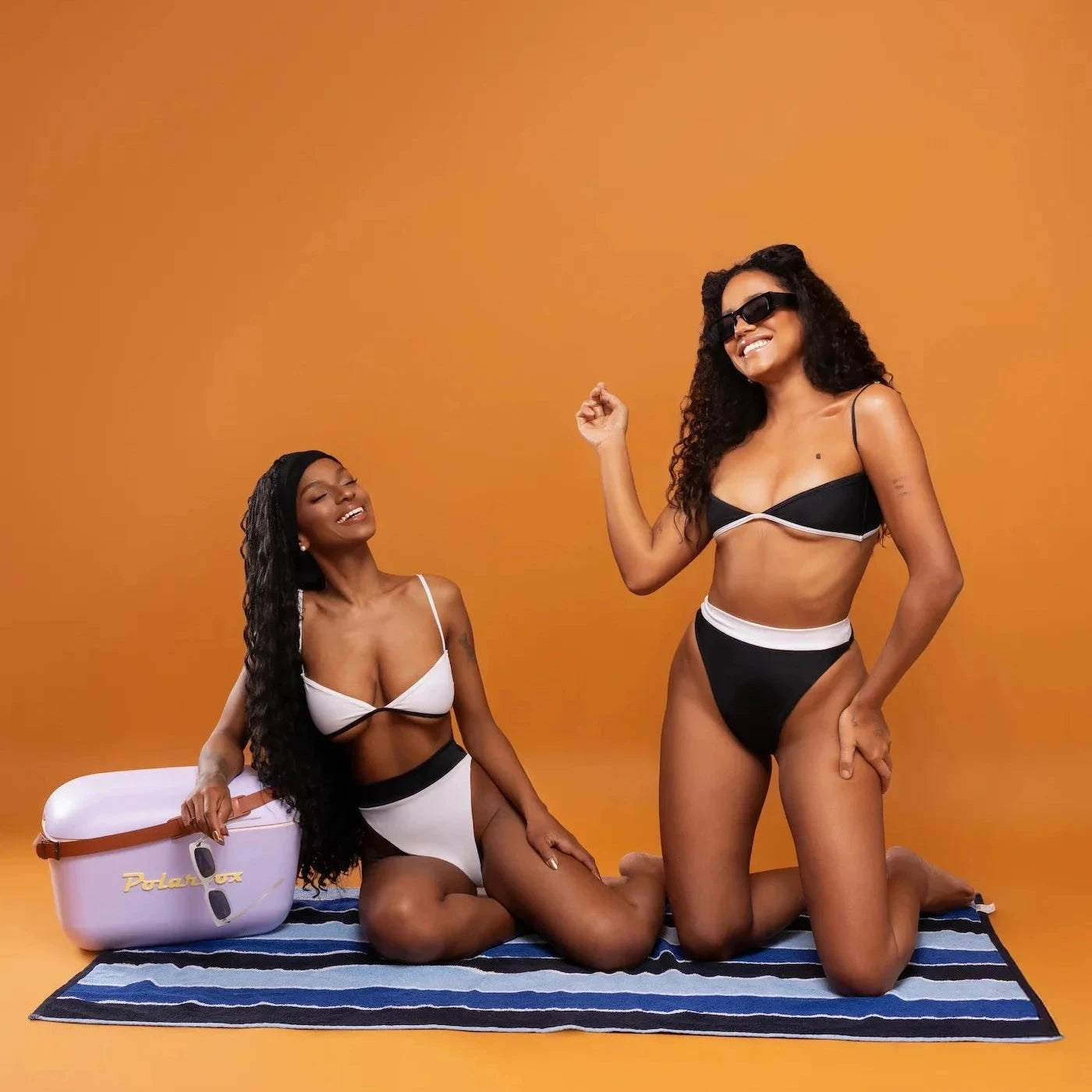 Two women in swimsuits posing on a striped towel against an orange background